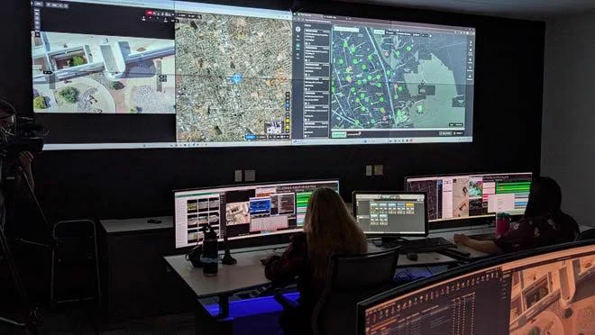 An operations room: pilots at consoles below a wall of live aerial feeds and maps