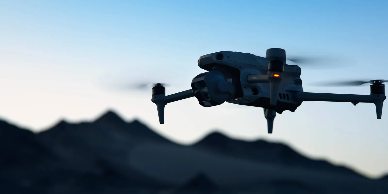 A Matrice 4 in flight at dusk, its multi sensor gimbal turned down toward the ground, a mountain ridge behind it