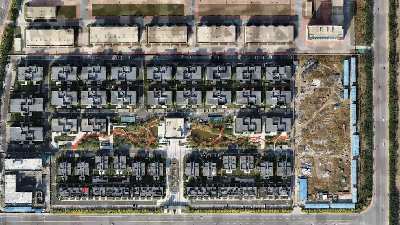 A top down aerial of a residential estate in daylight: roofs, streets, gardens and a construction plot, seen in ordinary colour