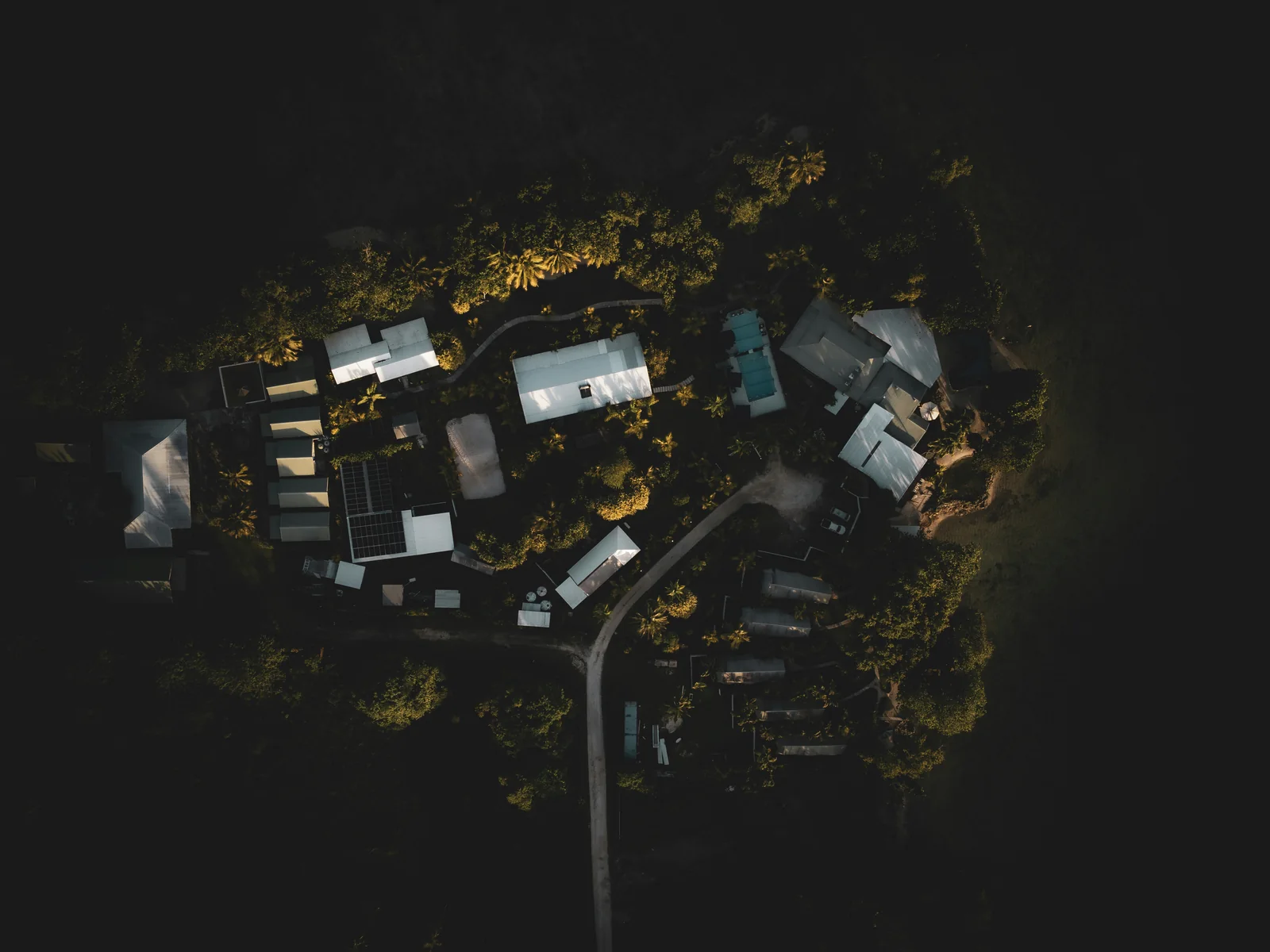 A rural compound seen straight down at night: sheds, cabins, water tanks, a solar array and one access track, with near black bush closing in on every side